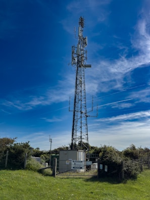 A tall communication tower surrounded by greenery under a vibrant blue sky with scattered clouds. The fenced area at the base contains technical equipment.