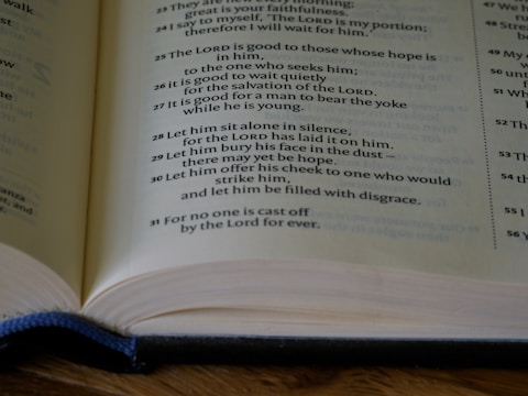 An open book with visible text on the pages, featuring religious scripture or verses. The lighting is soft, highlighting the words and pages. The text is printed in a standard font, with some phrases possibly emphasized in bold or different formatting.