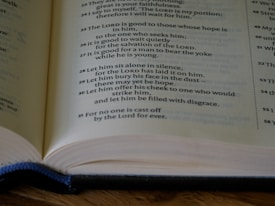 An open book with visible text on the pages, featuring religious scripture or verses. The lighting is soft, highlighting the words and pages. The text is printed in a standard font, with some phrases possibly emphasized in bold or different formatting.