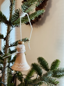 A wooden angel ornament with a serene expression hangs from a branch of an evergreen tree. The ornament features simple, hand-drawn details and is suspended by a piece of white string. The surrounding branches are lush and green, suggesting a festive, natural setting.