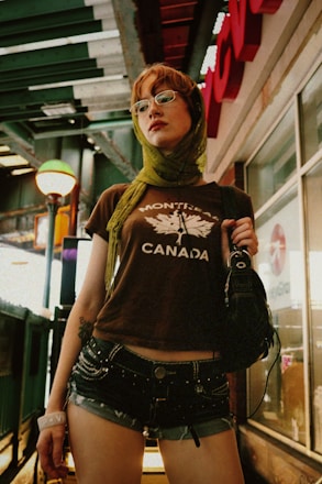A person wearing a t-shirt with the word 'Montreal' and a graphic, denim shorts, and a green scarf around their head and neck. They are standing under an outdoor structure with a large retail store sign in the background. The person is also wearing glasses and holding a bag, with a lamp post visible nearby.