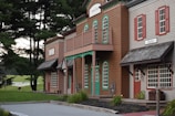 A rustic wooden building resembling an old Western town with signs indicating a hotel and barber shop. These buildings are set against a backdrop of tall, lush green trees and a distant grassy field with a white fence. A single alpaca or llama grazes in the background, adding a quaint farm-like charm to the scene.