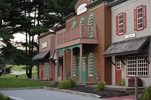 A rustic wooden building resembling an old Western town with signs indicating a hotel and barber shop. These buildings are set against a backdrop of tall, lush green trees and a distant grassy field with a white fence. A single alpaca or llama grazes in the background, adding a quaint farm-like charm to the scene.
