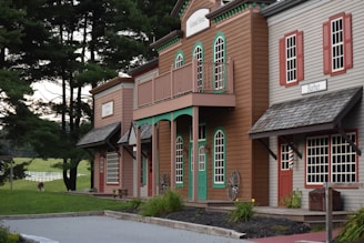 A rustic wooden building resembling an old Western town with signs indicating a hotel and barber shop. These buildings are set against a backdrop of tall, lush green trees and a distant grassy field with a white fence. A single alpaca or llama grazes in the background, adding a quaint farm-like charm to the scene.