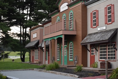 A rustic wooden building resembling an old Western town with signs indicating a hotel and barber shop. These buildings are set against a backdrop of tall, lush green trees and a distant grassy field with a white fence. A single alpaca or llama grazes in the background, adding a quaint farm-like charm to the scene.