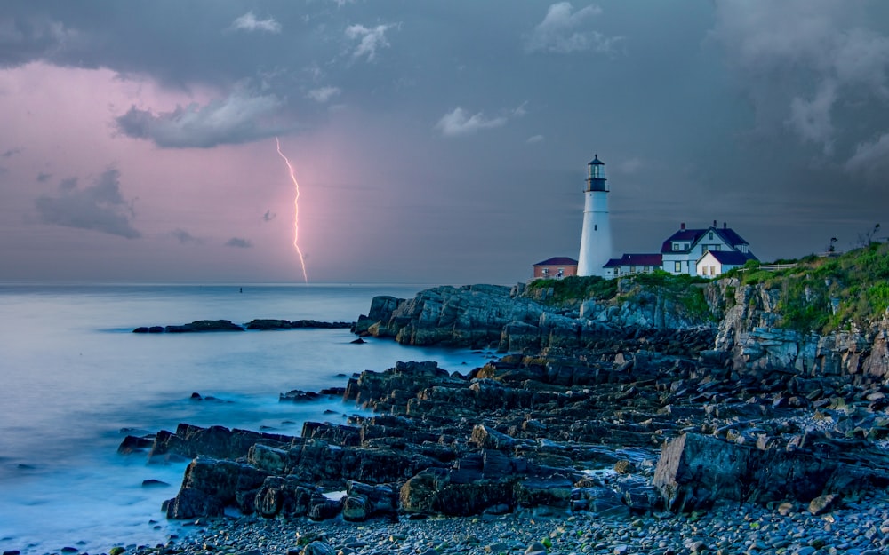 a lightning bolt strikes behind a lighthouse on a rocky shore