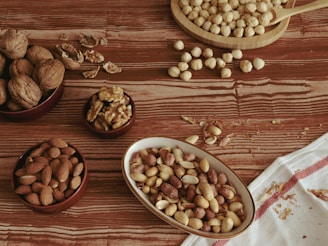 Various types of nuts are arranged on a wooden surface. There are bowls containing almonds, walnuts, hazelnuts, and peanuts. Some shelled nuts and crumbs are scattered around, adding a rustic touch. A white cloth with red stripes is placed partially on the surface.