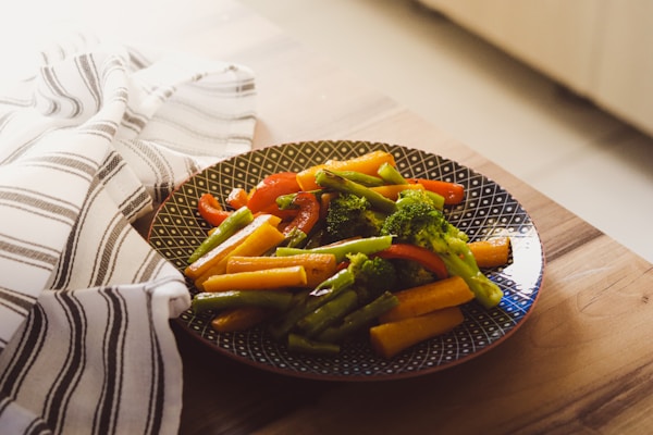 A plate of steamed vegetables, including broccoli, asparagus, carrots, and bell peppers, rests on a wooden table. The vegetables are arranged neatly on a patterned plate, and a striped dish towel lies beside it, adding a touch of rustic charm.