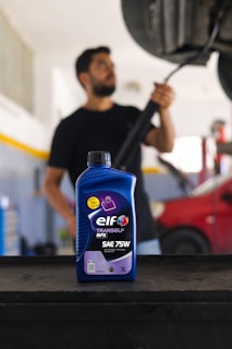 A bottle of ELF Tranself NFX SAE 75W gear oil is prominently displayed on a workbench, with a blurry person in the background holding a tool near a car undercarriage. The setting appears to be a garage or auto repair shop, featuring a red car and various equipment.