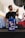 A bottle of ELF Tranself NFX SAE 75W gear oil is prominently displayed on a workbench, with a blurry person in the background holding a tool near a car undercarriage. The setting appears to be a garage or auto repair shop, featuring a red car and various equipment.