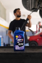 A bottle of ELF Tranself NFX SAE 75W gear oil is prominently displayed on a workbench, with a blurry person in the background holding a tool near a car undercarriage. The setting appears to be a garage or auto repair shop, featuring a red car and various equipment.