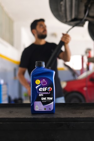 A bottle of ELF Tranself NFX SAE 75W gear oil is prominently displayed on a workbench, with a blurry person in the background holding a tool near a car undercarriage. The setting appears to be a garage or auto repair shop, featuring a red car and various equipment.