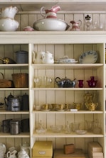 A wooden shelving unit filled with various vintage and ornate items including teapots, pitchers, cups, and decorative glassware. The shelves also display some metal teapots and woven baskets. There are delicate floral and gold patterns on some of the ceramics, adding an antique and elegant touch.