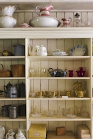 A wooden shelving unit filled with various vintage and ornate items including teapots, pitchers, cups, and decorative glassware. The shelves also display some metal teapots and woven baskets. There are delicate floral and gold patterns on some of the ceramics, adding an antique and elegant touch.