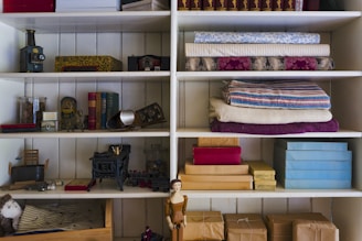 A set of wooden shelves filled with various vintage items, including old books, fabric rolls, wrapped packages, toy-like objects, a small mannequin doll, and decorative boxes. The fabric rolls are neatly stacked on the upper shelves, while boxes and books are arranged on the middle shelves. Several small decorative objects and toys are visible alongside these items.