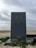 A large, gray vertical structure stands on a sandy beach. It has two faucets on the front and is marked with a large number 3 and a symbol resembling raindrops arranged in a triangle formation. The ground around the structure is wet and has some green plants scattered. The background features a cloudy sky and an expansive, mostly empty beach.