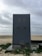 A large, gray vertical structure stands on a sandy beach. It has two faucets on the front and is marked with a large number 3 and a symbol resembling raindrops arranged in a triangle formation. The ground around the structure is wet and has some green plants scattered. The background features a cloudy sky and an expansive, mostly empty beach.