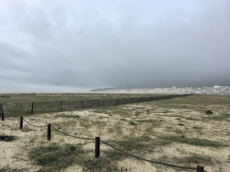 A vast, sandy landscape under an overcast sky with heavy clouds. A long, wooden fence stretches into the distance, dividing the sparsely vegetated sand from a town with clustered buildings visible in the background.