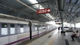 A modern metro train station with a train on the platform. The station is spacious and covered by a metal roof. There are a few people on the platform, some seated and others walking. A prominent sign above the platform reads 'Emergency Train Stop' in English and another language.