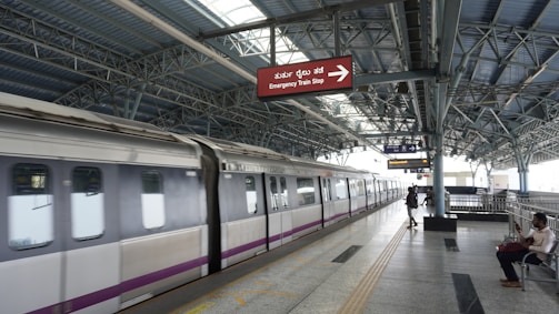 A modern metro train station with a train on the platform. The station is spacious and covered by a metal roof. There are a few people on the platform, some seated and others walking. A prominent sign above the platform reads 'Emergency Train Stop' in English and another language.