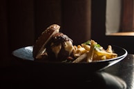 A gourmet burger with melted cheese and caramelized onions is served on a black plate alongside a pile of golden, crispy French fries. The lighting is dramatic, highlighting the textures of the sesame seed bun and the fries. A small window in the background adds a warm glow to the scene.