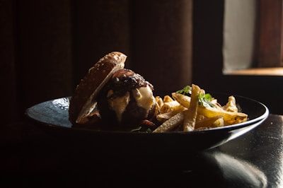 A gourmet burger with melted cheese and caramelized onions is served on a black plate alongside a pile of golden, crispy French fries. The lighting is dramatic, highlighting the textures of the sesame seed bun and the fries. A small window in the background adds a warm glow to the scene.