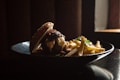 A gourmet burger with melted cheese and caramelized onions is served on a black plate alongside a pile of golden, crispy French fries. The lighting is dramatic, highlighting the textures of the sesame seed bun and the fries. A small window in the background adds a warm glow to the scene.