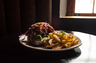 A plate of food featuring a sandwich loaded with various toppings, including purple cabbage and leafy greens, is accompanied by a generous serving of golden-brown fries. The setting appears to have minimal lighting, with a warm, moody atmosphere created by the dark background and a window on the side letting in some light.