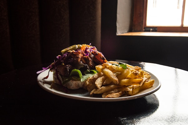 A plate of food featuring a sandwich loaded with various toppings, including purple cabbage and leafy greens, is accompanied by a generous serving of golden-brown fries. The setting appears to have minimal lighting, with a warm, moody atmosphere created by the dark background and a window on the side letting in some light.