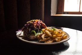 A plate of food featuring a sandwich loaded with various toppings, including purple cabbage and leafy greens, is accompanied by a generous serving of golden-brown fries. The setting appears to have minimal lighting, with a warm, moody atmosphere created by the dark background and a window on the side letting in some light.