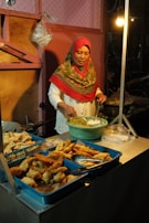 A woman wearing a headscarf is preparing and serving fried snacks from multiple trays at a food stall. She is using a spoon to mix ingredients in a green bowl. The setting is dimly lit, suggesting an evening or night market.
