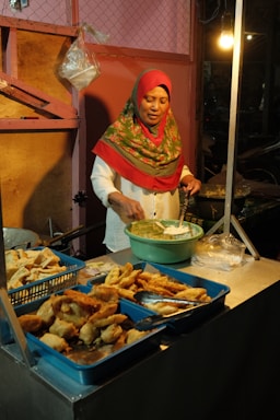 A woman wearing a headscarf is preparing and serving fried snacks from multiple trays at a food stall. She is using a spoon to mix ingredients in a green bowl. The setting is dimly lit, suggesting an evening or night market.