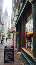 A bustling city street scene featuring a restaurant with neon signage displaying 'Olde City' in the foreground. The entrance is flanked by a board listing menus and specials. There are tall buildings along the street, with a view of a towering structure in the background. Pedestrians are walking on the sidewalk, and the atmosphere suggests a typical busy urban area.