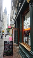 A bustling city street scene featuring a restaurant with neon signage displaying 'Olde City' in the foreground. The entrance is flanked by a board listing menus and specials. There are tall buildings along the street, with a view of a towering structure in the background. Pedestrians are walking on the sidewalk, and the atmosphere suggests a typical busy urban area.