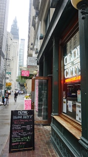 A bustling city street scene featuring a restaurant with neon signage displaying 'Olde City' in the foreground. The entrance is flanked by a board listing menus and specials. There are tall buildings along the street, with a view of a towering structure in the background. Pedestrians are walking on the sidewalk, and the atmosphere suggests a typical busy urban area.