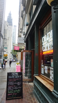 A bustling city street scene featuring a restaurant with neon signage displaying 'Olde City' in the foreground. The entrance is flanked by a board listing menus and specials. There are tall buildings along the street, with a view of a towering structure in the background. Pedestrians are walking on the sidewalk, and the atmosphere suggests a typical busy urban area.