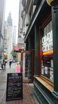 A bustling city street scene featuring a restaurant with neon signage displaying 'Olde City' in the foreground. The entrance is flanked by a board listing menus and specials. There are tall buildings along the street, with a view of a towering structure in the background. Pedestrians are walking on the sidewalk, and the atmosphere suggests a typical busy urban area.