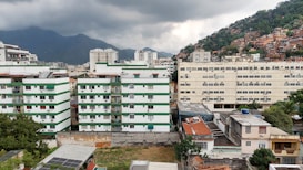 Several residential buildings are seen with a cloudy sky. The buildings are primarily white with green accents. In the background, there are mountains and a densely populated hillside with many houses. The foreground includes some greenery and smaller structures.