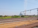 Railway tracks stretch across the foreground amidst a vast, open landscape with green fields. In the background, a large industrial facility with tall chimneys emitting smoke is visible. Electricity pylons and cables are also present, cutting across the sky, which is clear and blue.