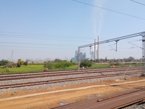 Railway tracks stretch across the foreground amidst a vast, open landscape with green fields. In the background, a large industrial facility with tall chimneys emitting smoke is visible. Electricity pylons and cables are also present, cutting across the sky, which is clear and blue.