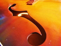 A close-up view of an f-hole on a string instrument, showcasing the wood grain and vibrant reddish-brown tones. The lighting highlights the smooth finish and the elegant curves of the design.
