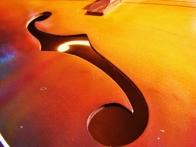 A close-up view of an f-hole on a string instrument, showcasing the wood grain and vibrant reddish-brown tones. The lighting highlights the smooth finish and the elegant curves of the design.