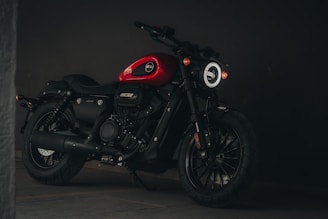 A sleek black motorcycle with a red fuel tank stands in a dimly lit environment. The motorcycle features a rugged build, thick tires, and a modern round headlight. Its design elements suggest a blend of vintage and contemporary styling.