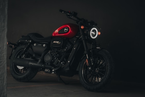 A sleek black motorcycle with a red fuel tank stands in a dimly lit environment. The motorcycle features a rugged build, thick tires, and a modern round headlight. Its design elements suggest a blend of vintage and contemporary styling.