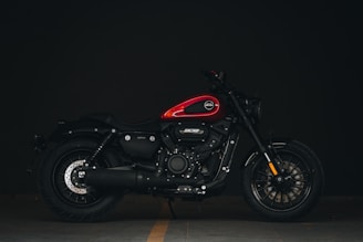 A sleek, modern motorcycle with a red fuel tank and black body is parked against a dark background. The design features robust tires, a prominent engine cover, and detailed metalwork with a focus on the front and rear wheels.