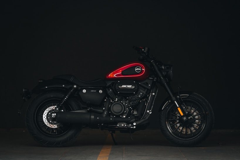 A sleek, modern motorcycle with a red fuel tank and black body is parked against a dark background. The design features robust tires, a prominent engine cover, and detailed metalwork with a focus on the front and rear wheels.