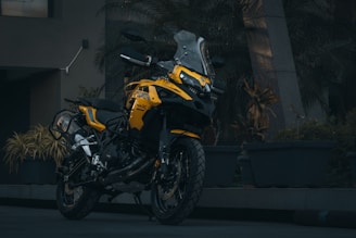 A sleek yellow motorcycle is parked on a paved surface, surrounded by large potted plants. The motorcycle has a modern design with prominent features like a windshield and angular bodywork. The setting appears to be outdoors, possibly near a building entrance, with dim lighting suggesting a moody ambiance.