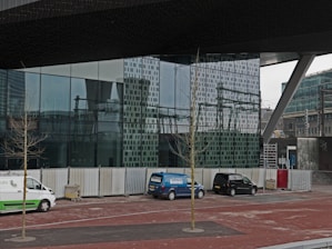 A modern architectural setting with a large glass facade reflecting the surrounding urban environment. Several vehicles, including a white and green van, a blue van, and a black car, are parked along the sidewalk. Leafless trees are planted along the pavement, and temporary fencing lines the area, suggesting construction or renovation activity.
