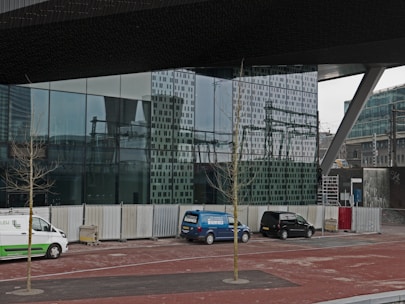 A modern architectural setting with a large glass facade reflecting the surrounding urban environment. Several vehicles, including a white and green van, a blue van, and a black car, are parked along the sidewalk. Leafless trees are planted along the pavement, and temporary fencing lines the area, suggesting construction or renovation activity.