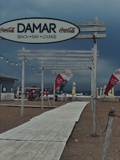 A beach bar and lounge with wooden structures and Coca-Cola branding. The area appears empty, with a pathway leading down to a sandy beach. Several flags and chairs are present near the shoreline.